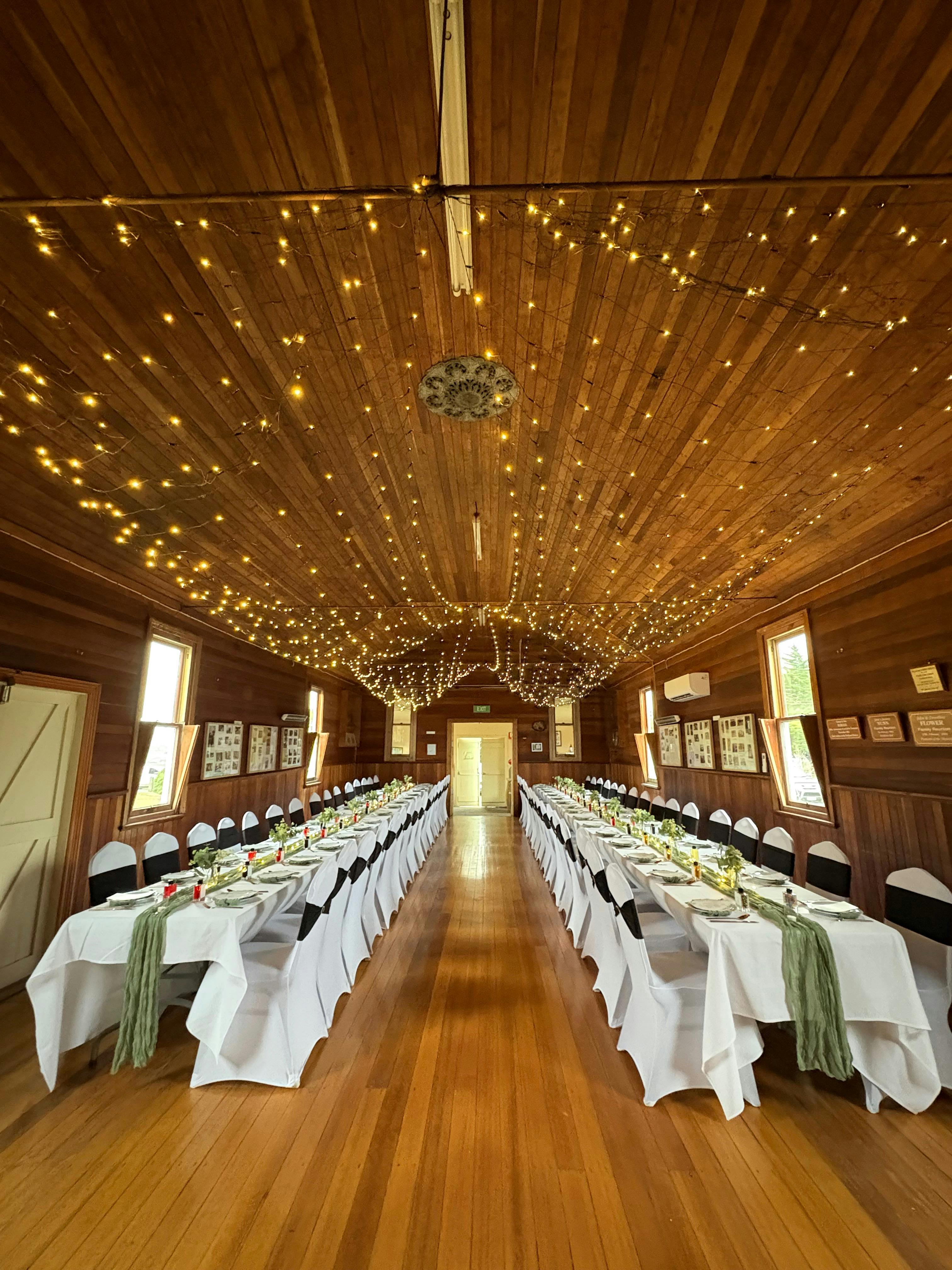 2 long tables set for banquet dinner in wooden hall with decorative lighting