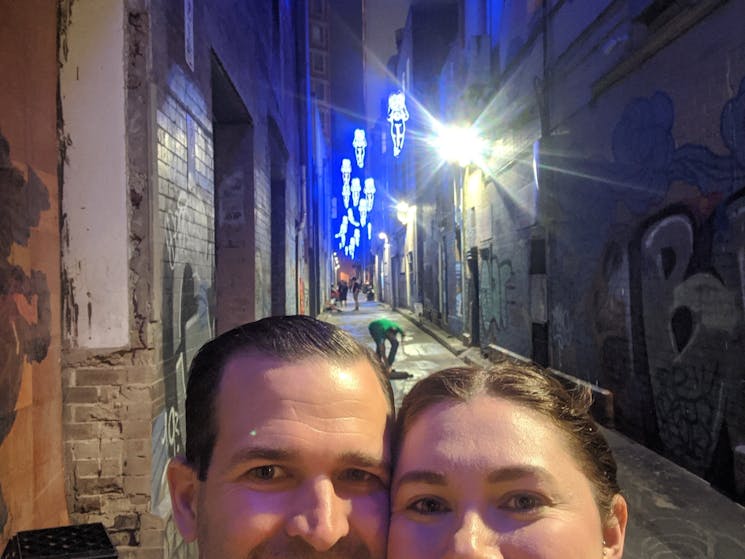 A couple posing in an alleyway with blue-lit art installation
