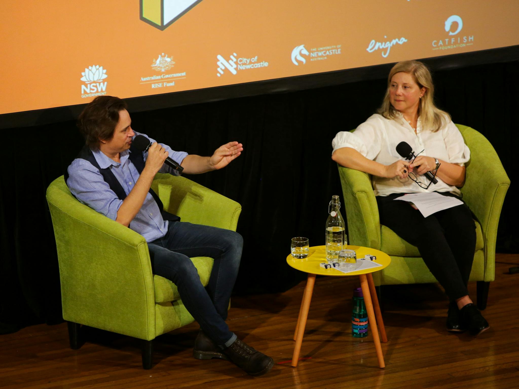 Author Trent Dalton is holding a microphone and gesturing to interviewer Rosemarie Milsom.