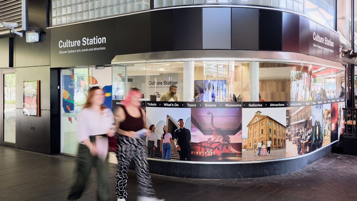 Two women walking past an information hub for Sydney's arts, culture and events
