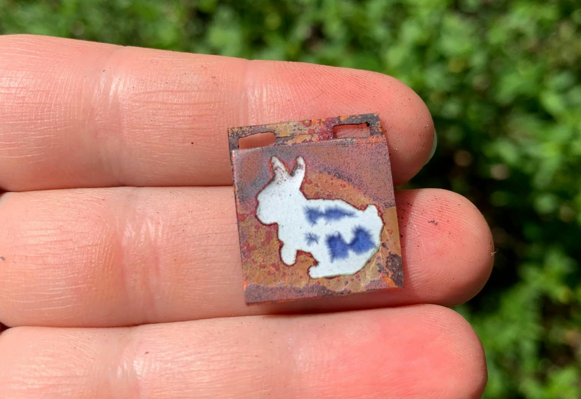 a photo of a copper pendant being held in the hand. The copper pendant has a glass enamel rabbit