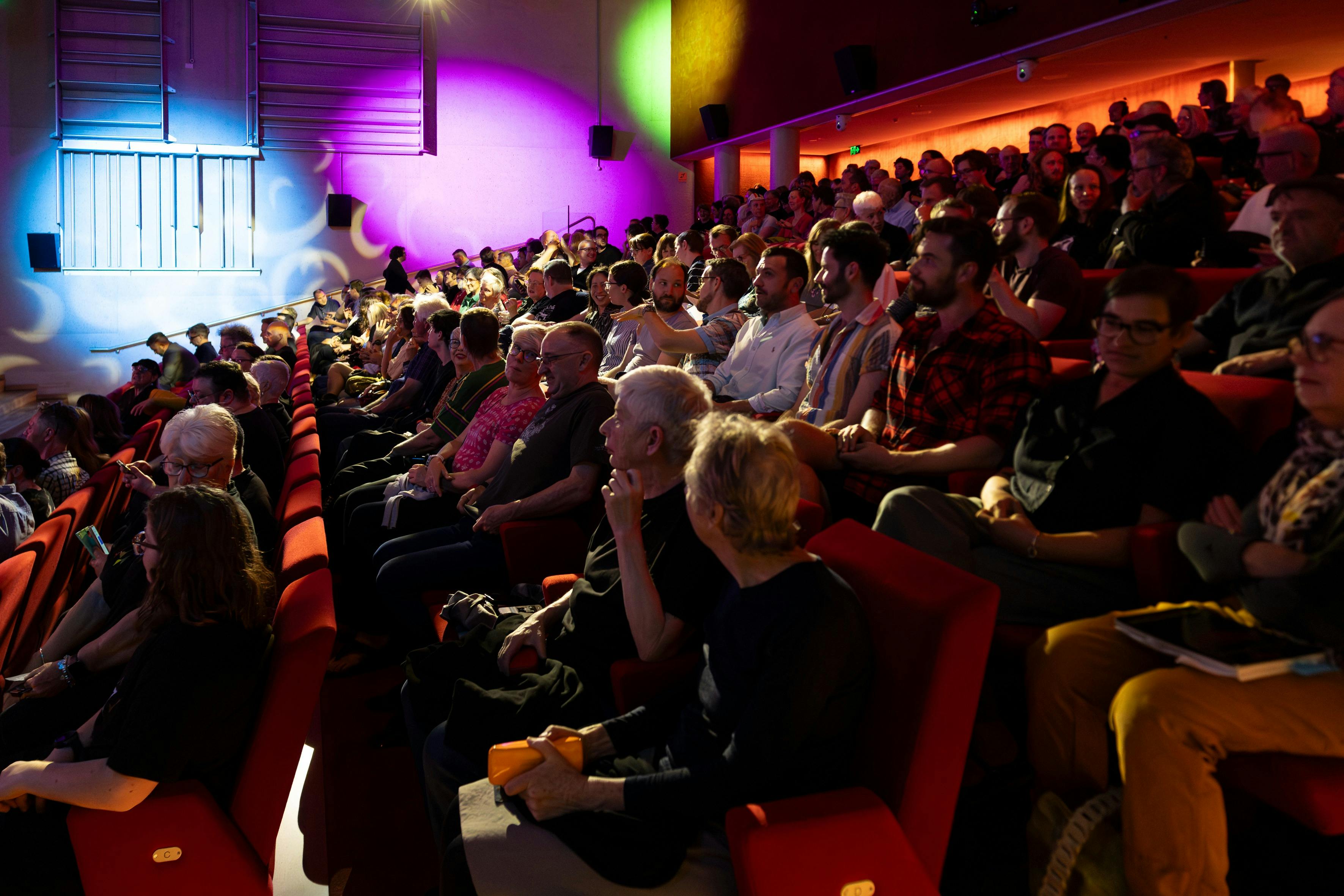 A photograph of a seated theatre audience talking with coloured lights in the background