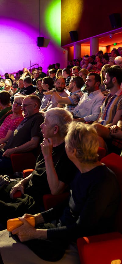 A photograph of a seated theatre audience talking with coloured lights in the background
