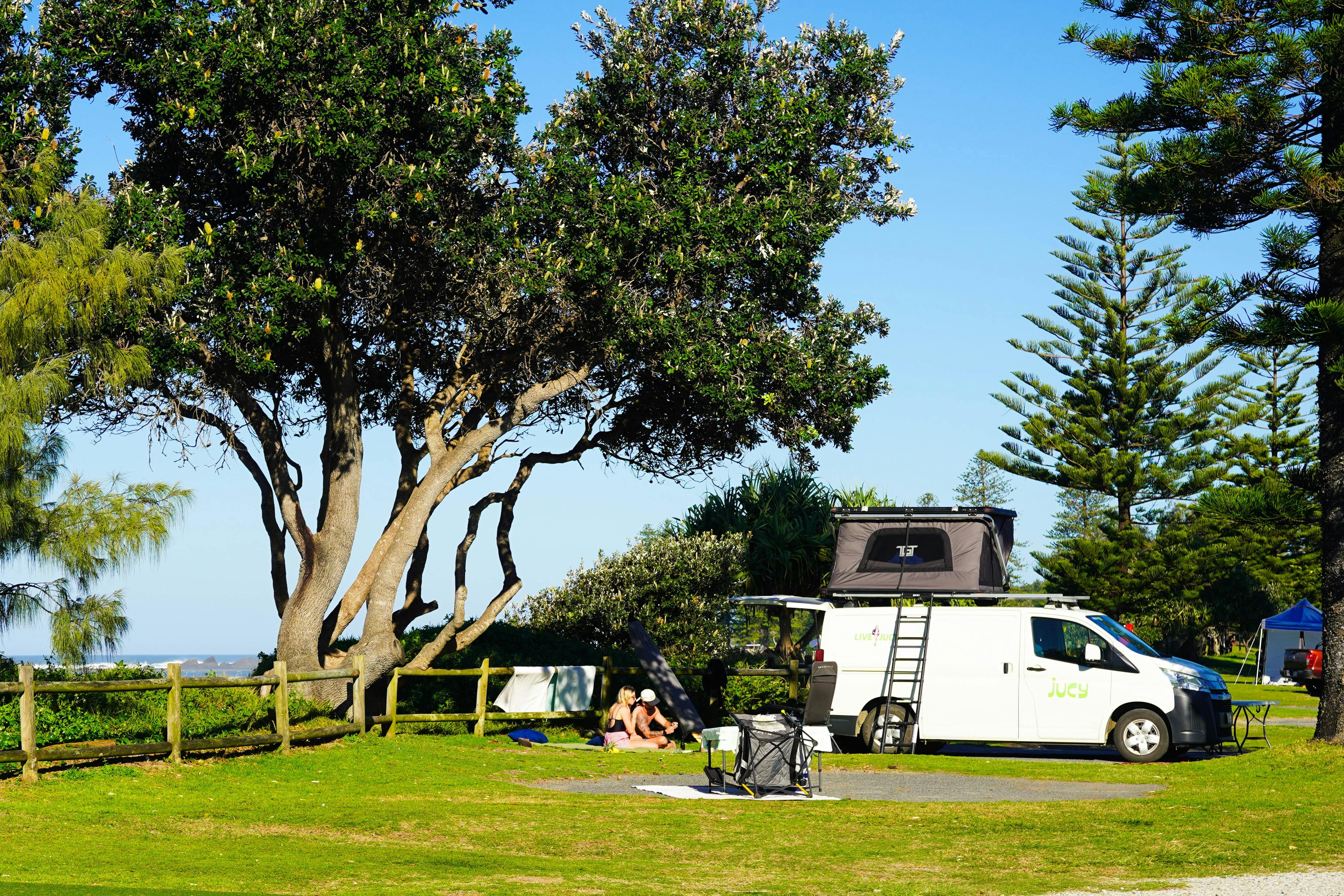Ein Wohnmobil mit einem auf einer grünen Wiese aufgebauten Dachzelt, daneben entspannen sich Camper auf einer Picknickdecke.