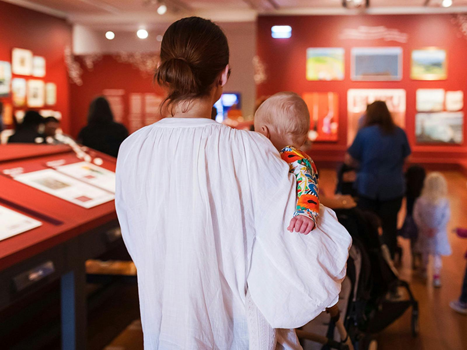 A woman holds a baby in a red-walled gallery space.