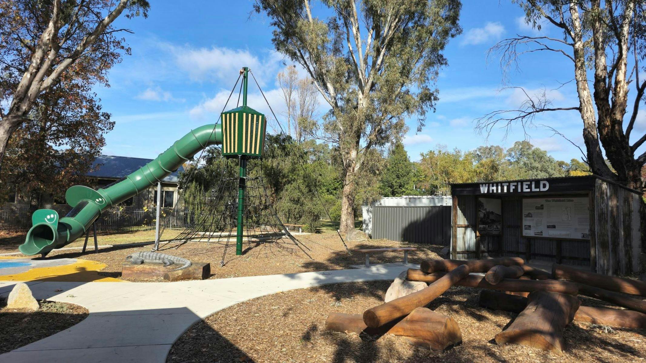 Concrete path going through bark spaces. Large green slide over a climbing net. Shed in background