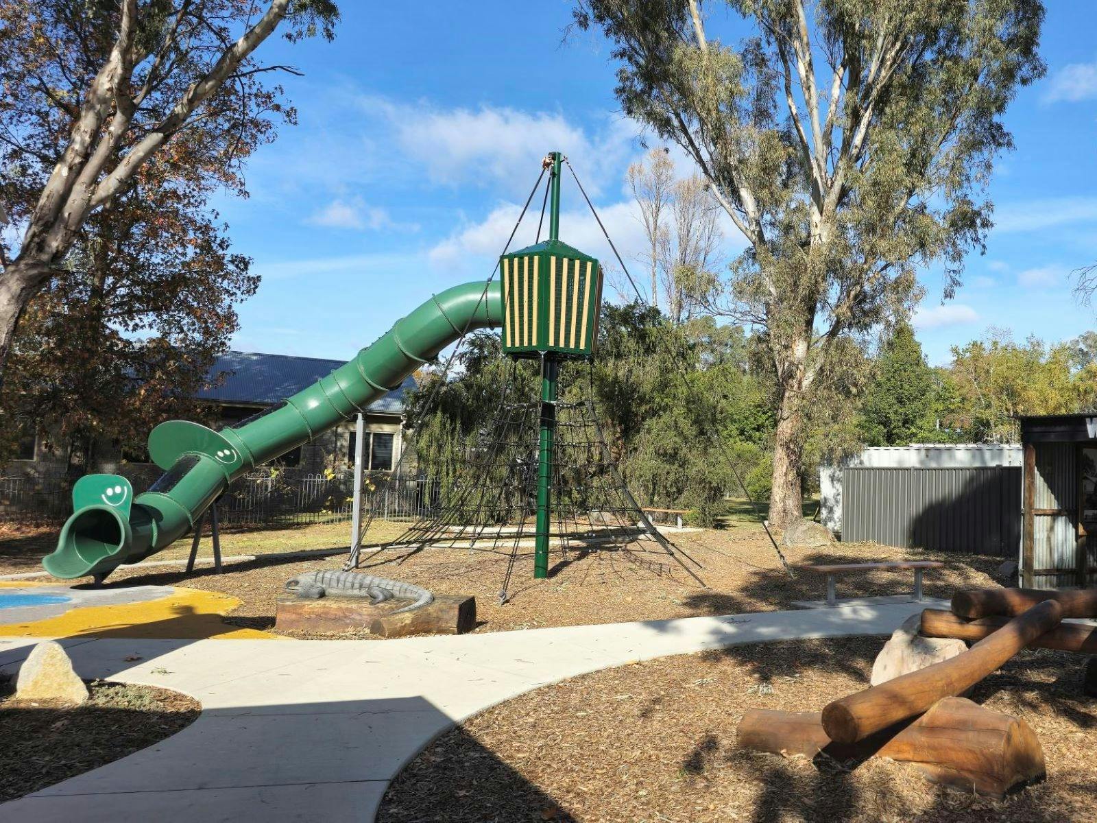 Concrete path going through bark spaces. Large green slide over a climbing net. Shed in background