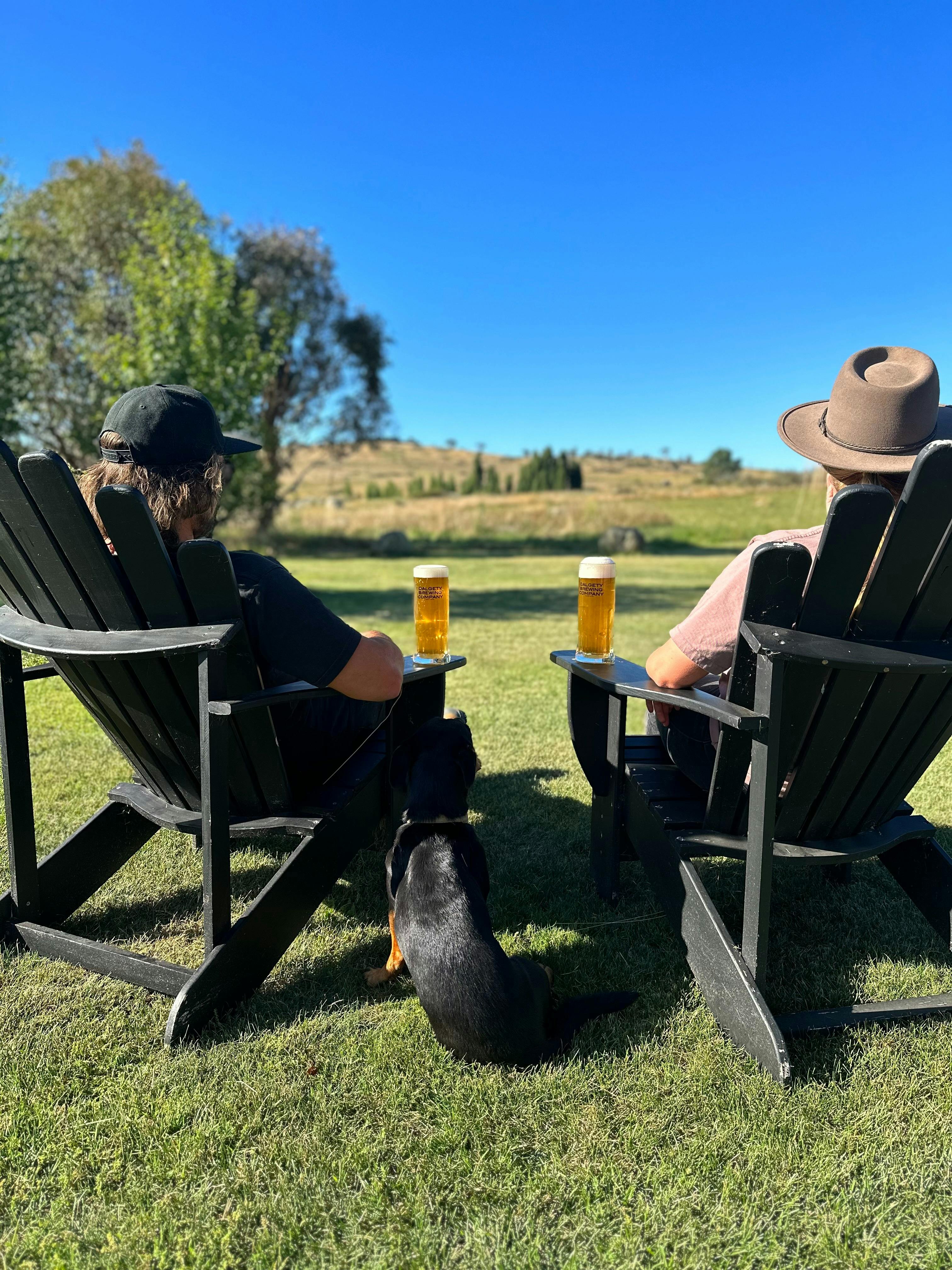 A couple and their dog sitting on two deck chairs with beers, looking out at the views
