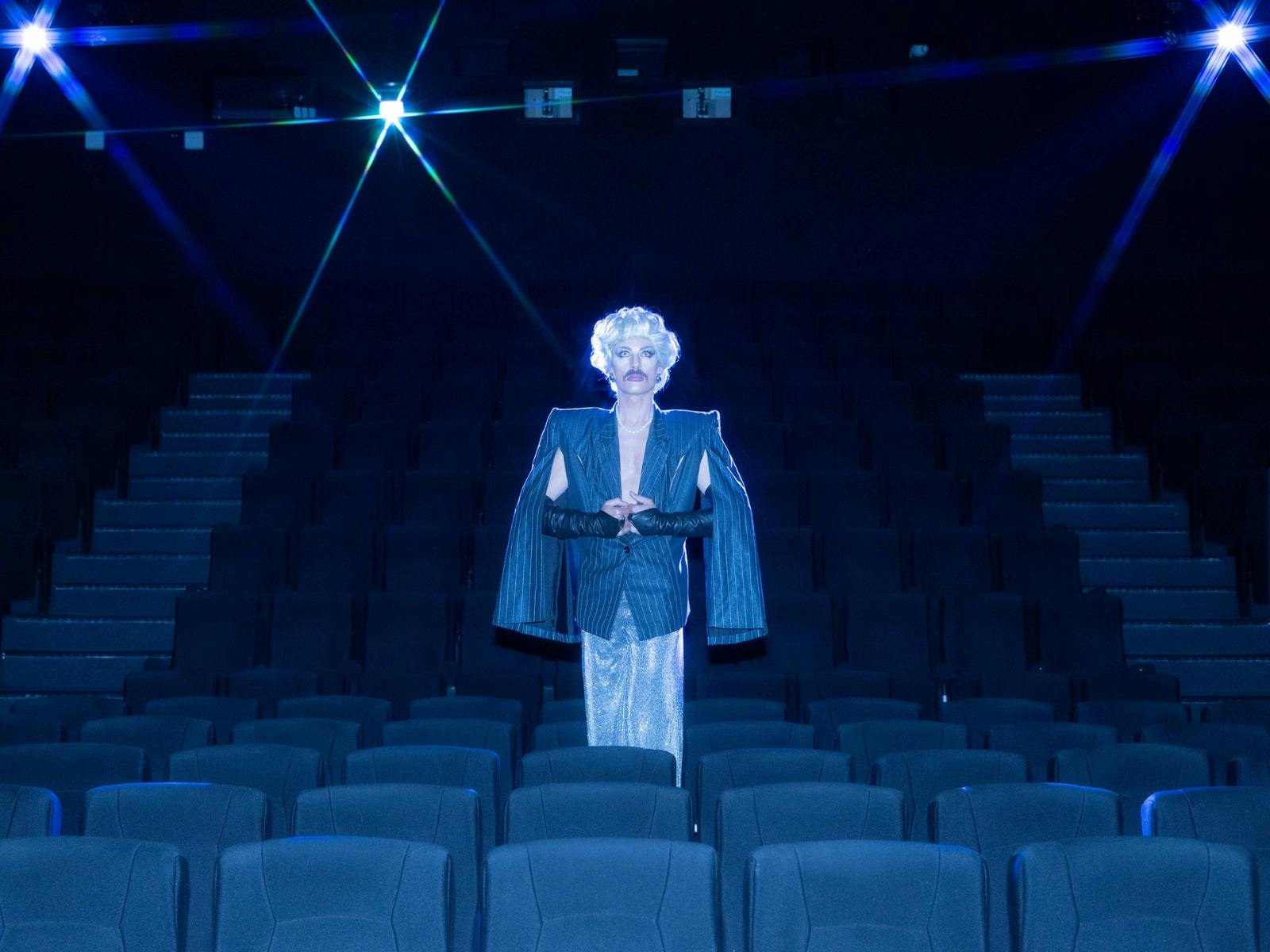 Drag artist stands in the middle of an empty cinema wearing an overcoat and blonde wig