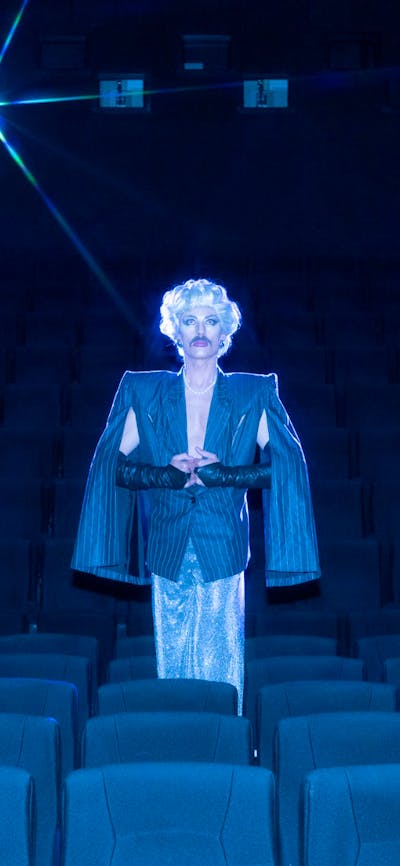 Drag artist stands in the middle of an empty cinema wearing an overcoat and blonde wig