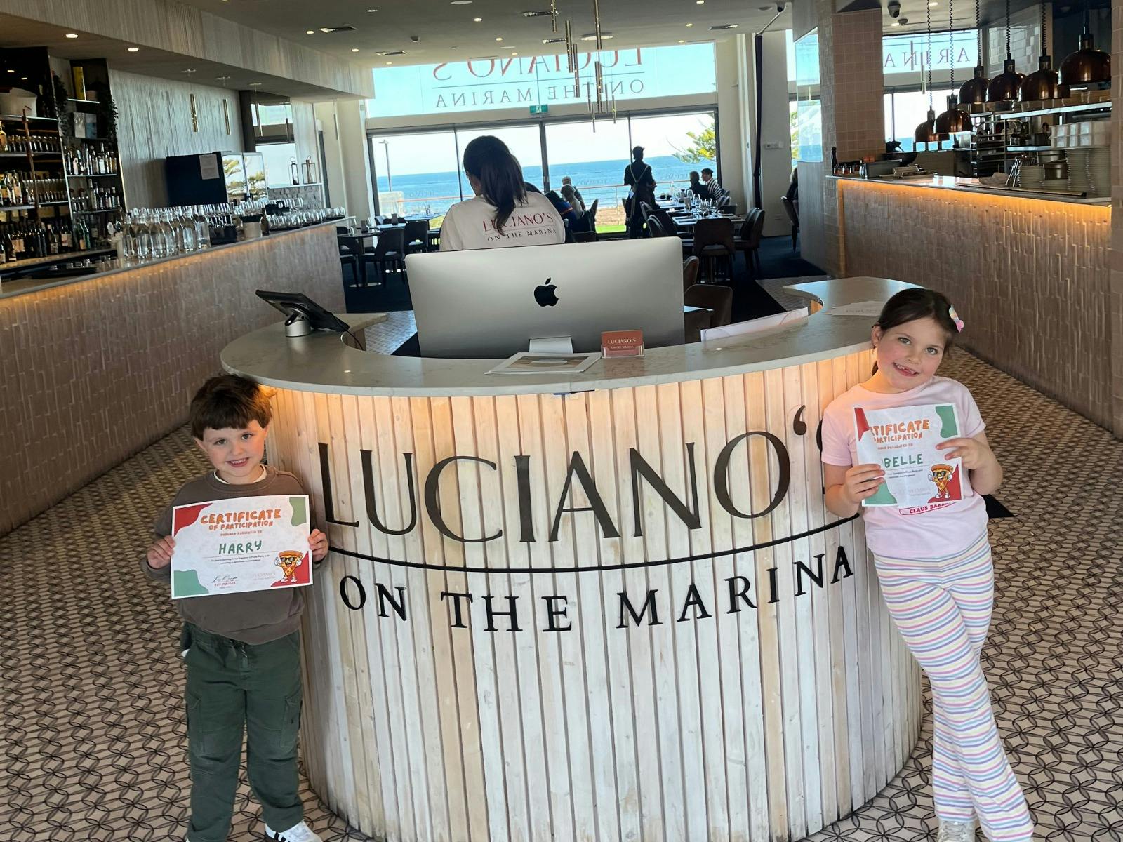 Two kids holding up certificates in front of Luciano's restaurant welcome desk sign
