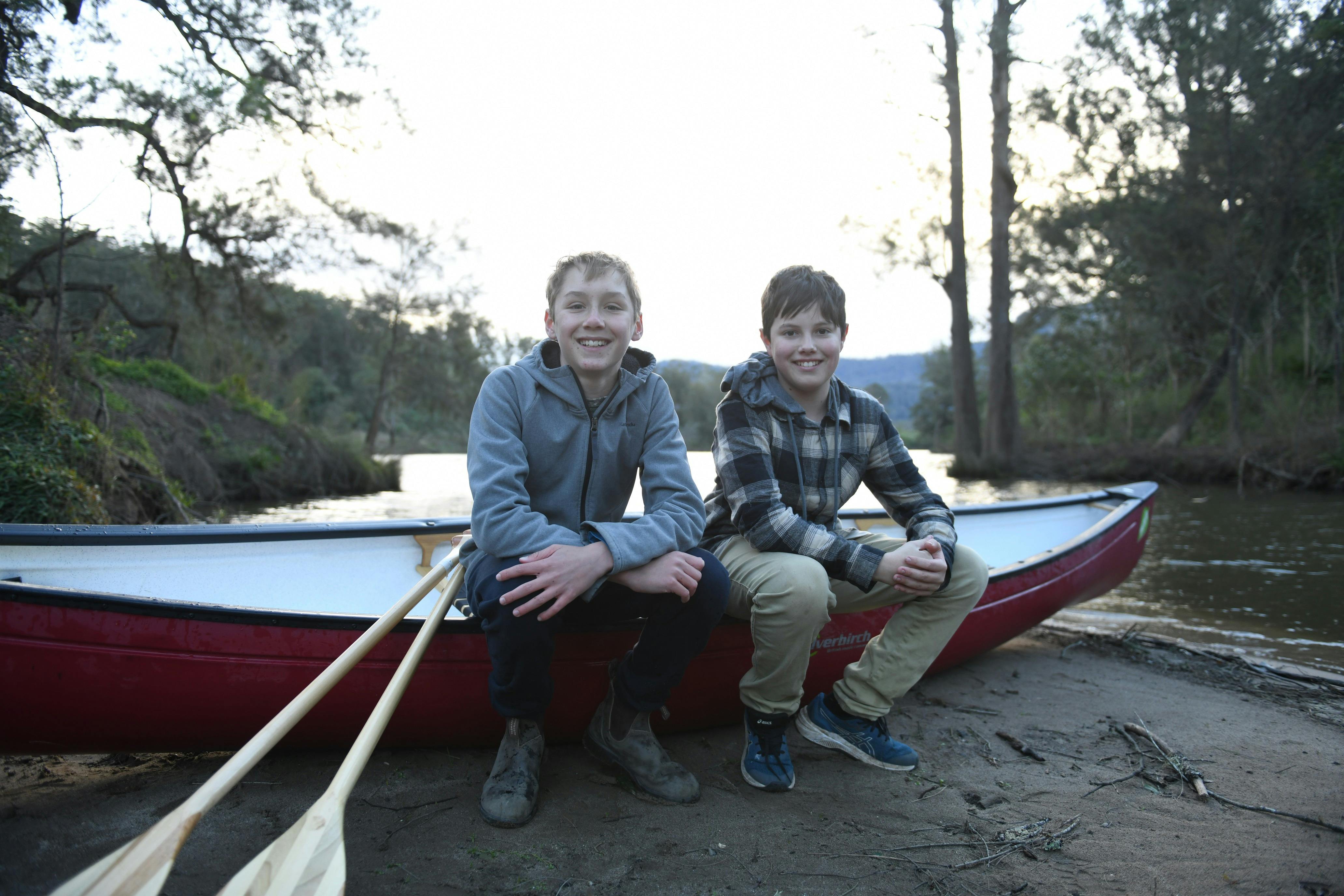 2 boys sitting on a canoe on land with a lake in the background