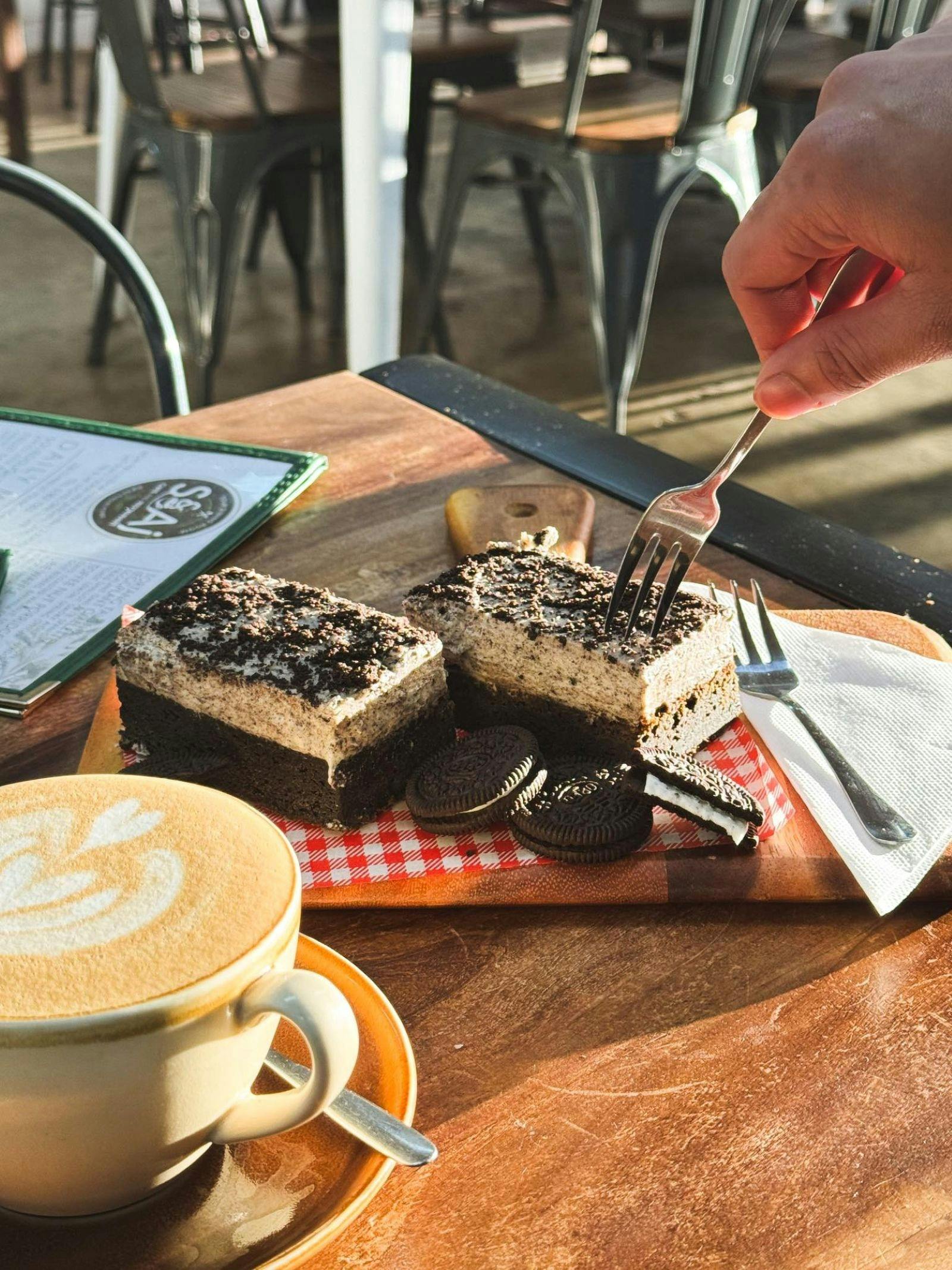 Serving of oreo brownie slice with someone pressing into it with a fork. Coffee on the side.