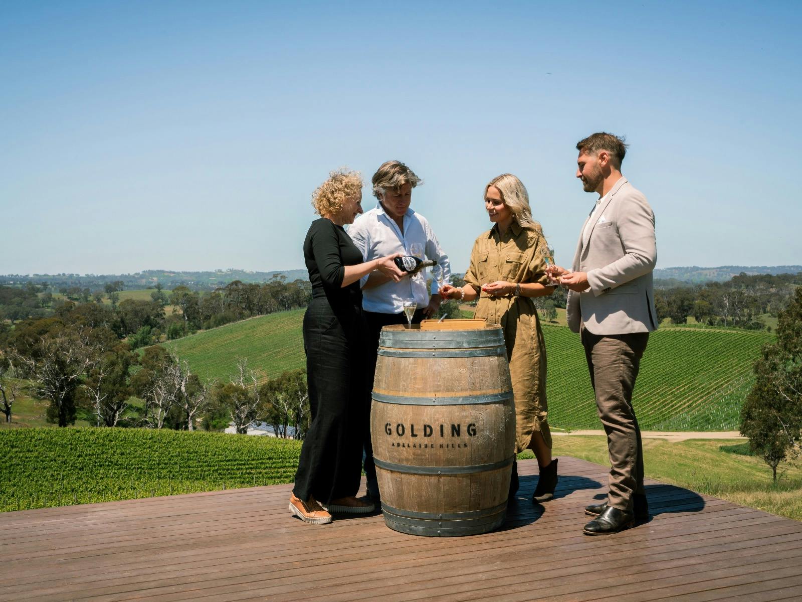 Four people stand around a barrel on a timber deck enjoying sparkling wine with a vineyard view.