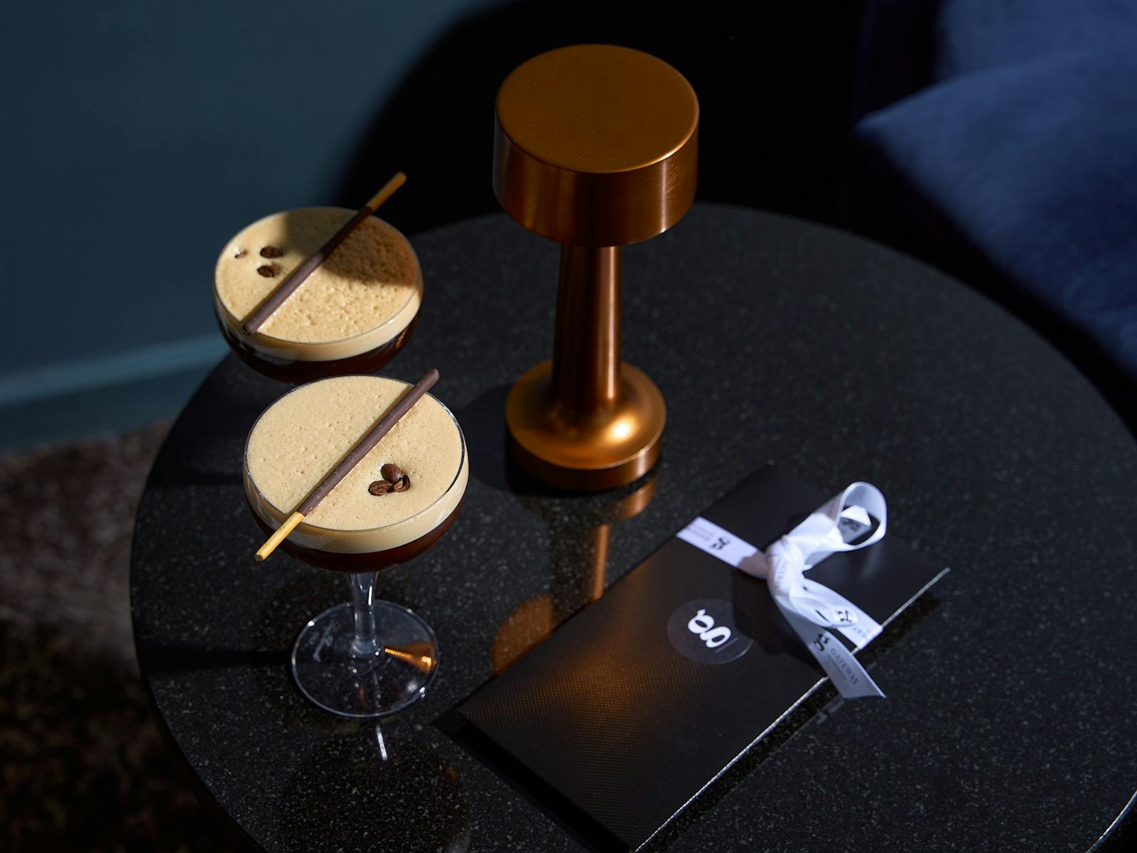 Two cocktails presented in candle lit bar with blue velvet sofas and gift with ribbon