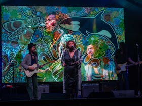 Singer Emma Donovan sings on stage with a guitarist in front of a backdrop of Aboriginal art
