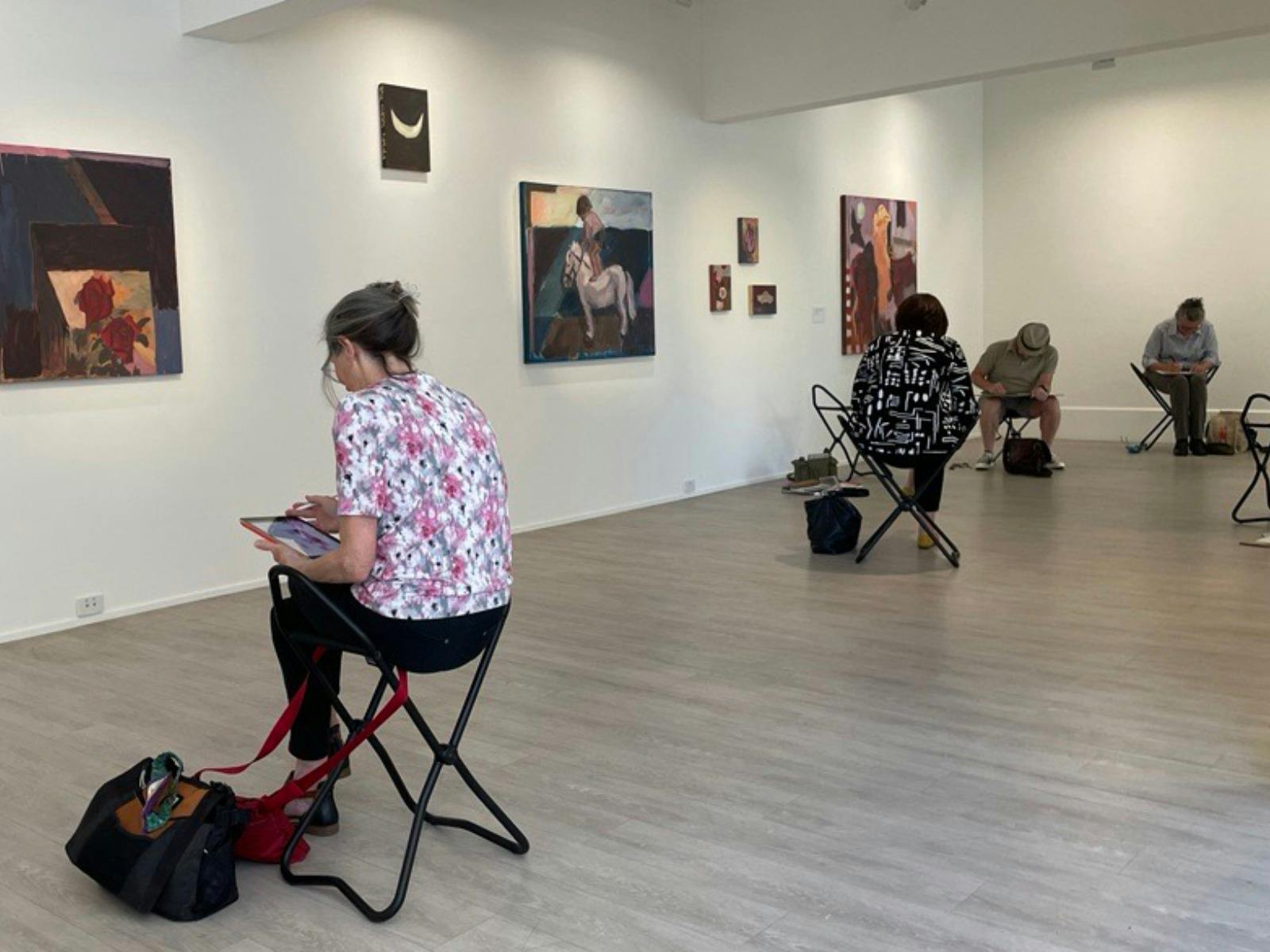 Four people sitting on pause stools in an exhibition and sketching on paper