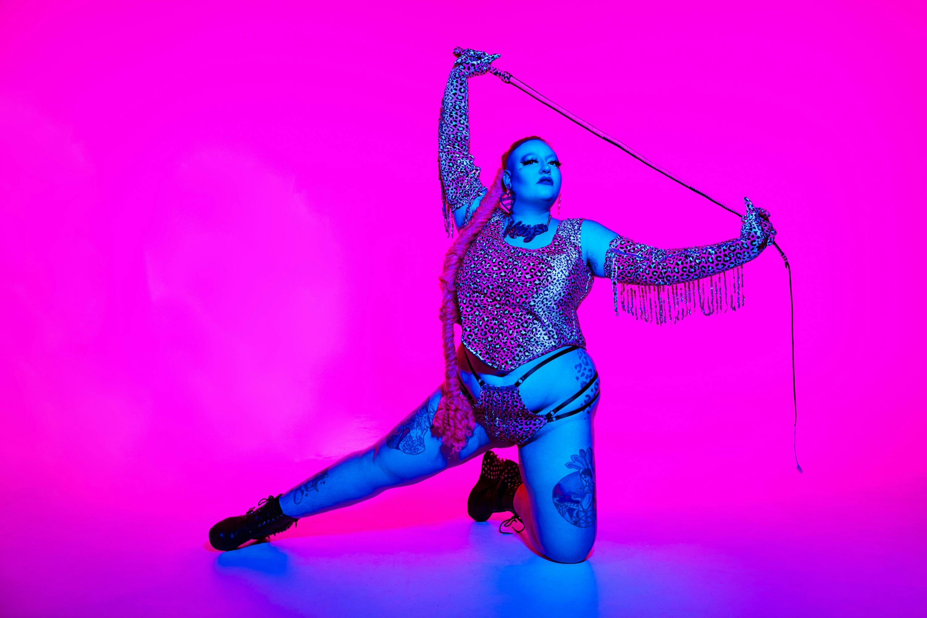 A curvy, drag performer with long pink hair appears on a pink and blue background.
