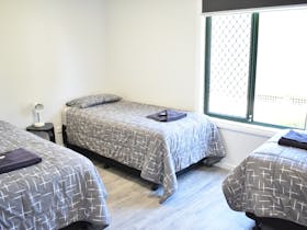 Bedroom with 3 single beds, grey patterned bedding, bedside table with a lamp and large window.