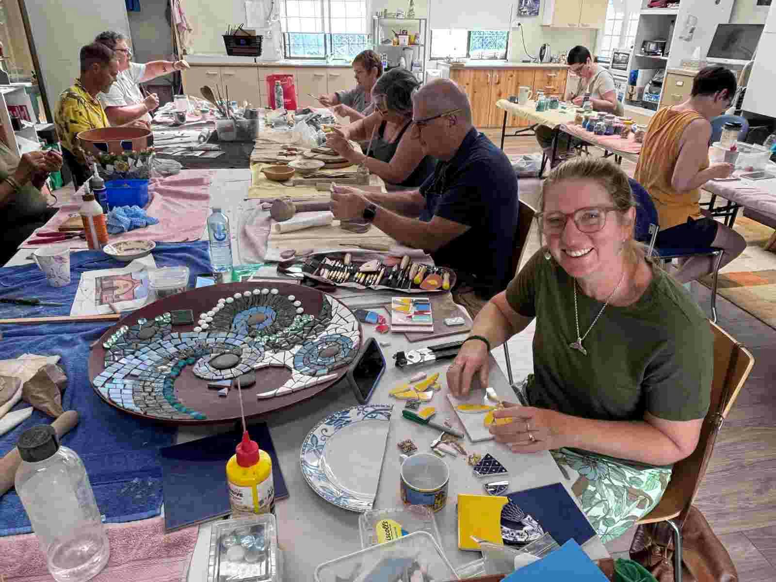 a group of peooel sitting at tables, working on their mosaics pieces