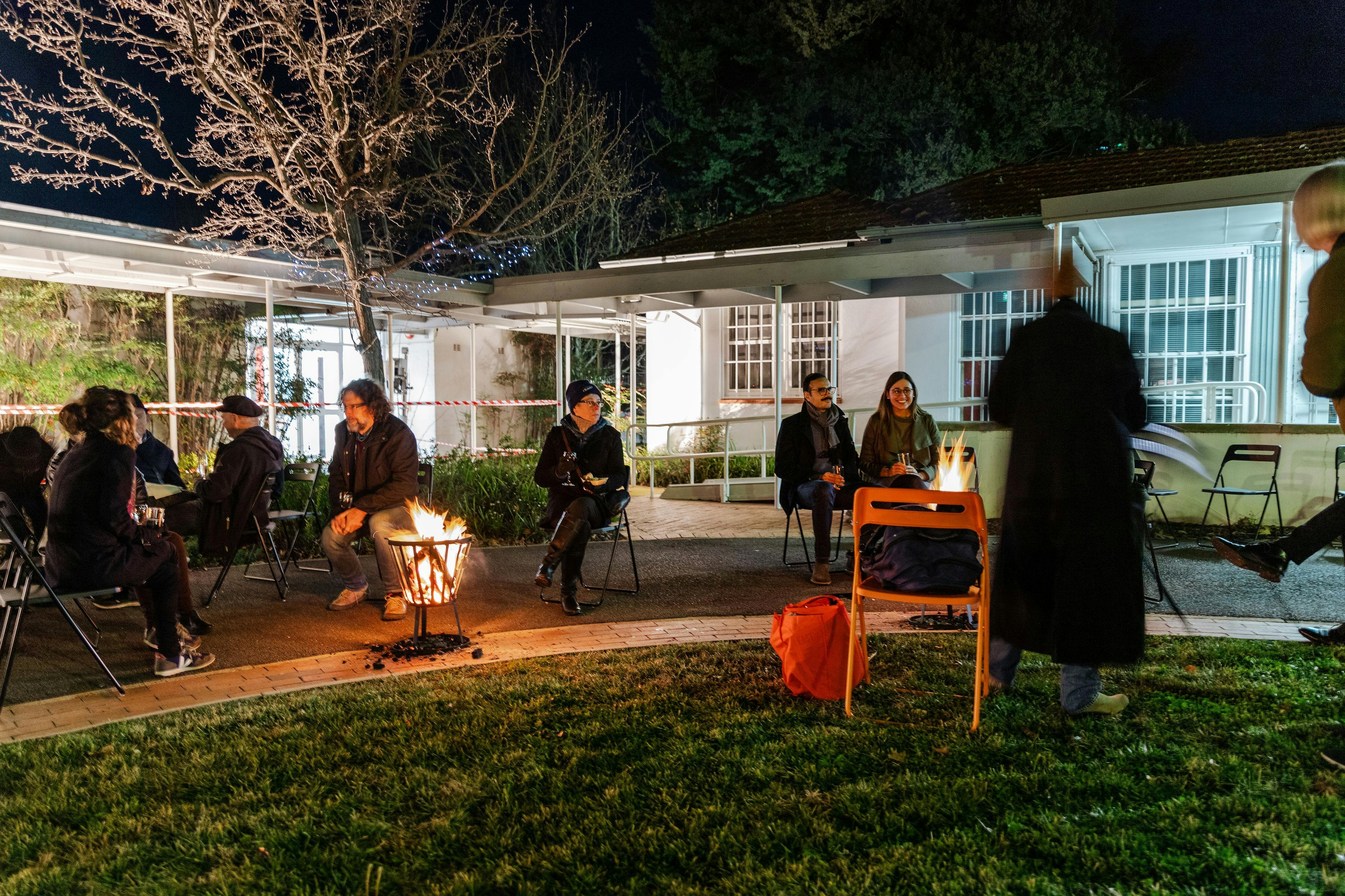 a group of people seated around a fire in a garden, outside a gallery.