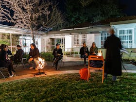 a group of people seated around a fire in a garden, outside a gallery.