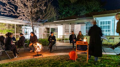 a group of people seated around a fire in a garden, outside a gallery.