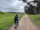 Cyclist on gravel road green grass, Large Gum trees on right grey cloudy sky
