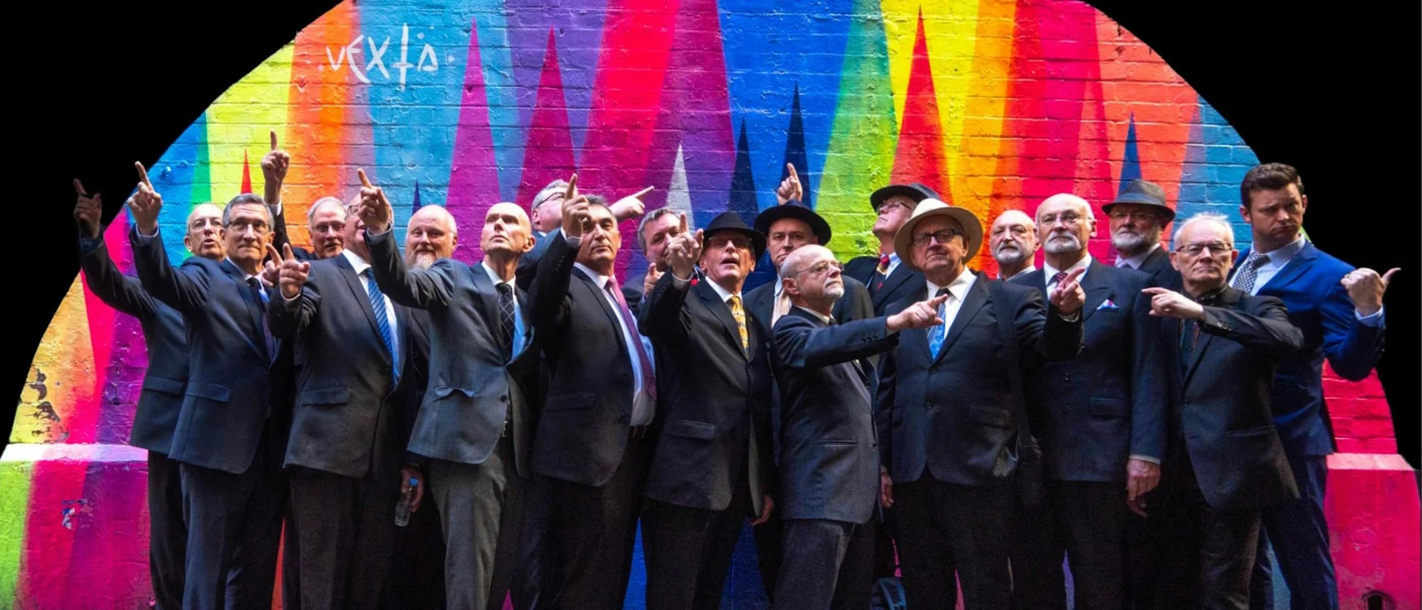 The singers of Men in Suits wearing business suits standing in front of a rainbow-coloured wall
