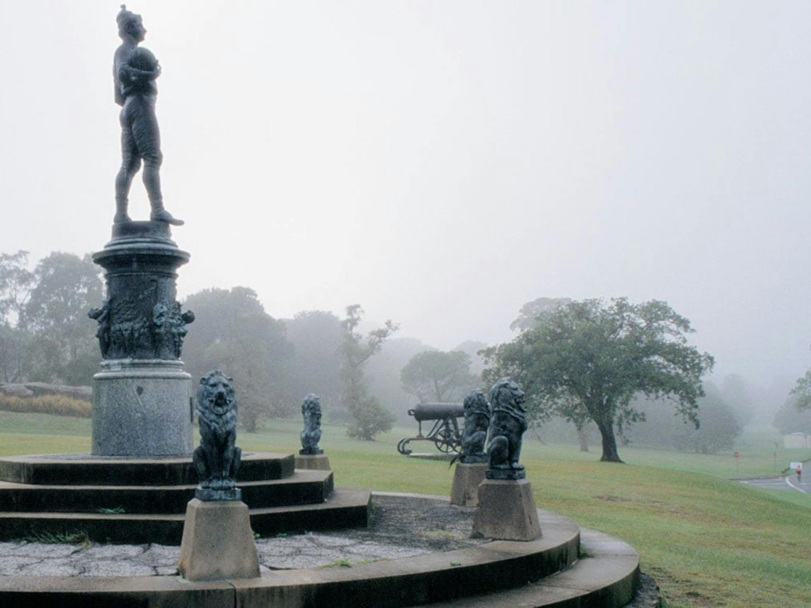 Bronze statue stands tall on a plynth. Below, figures surround. Mist covers trees in the background.