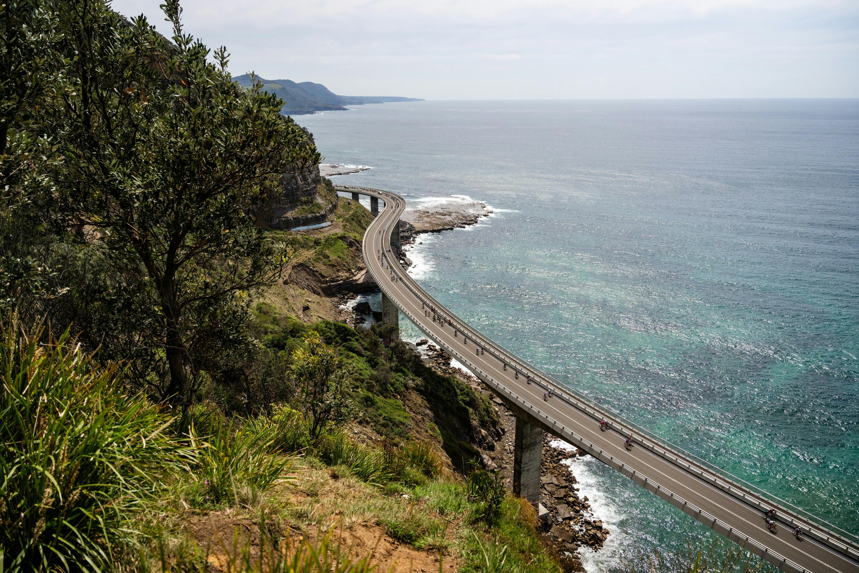 Bike riders crossing Sea Cliff Bridge, Ocean and Mountain views.