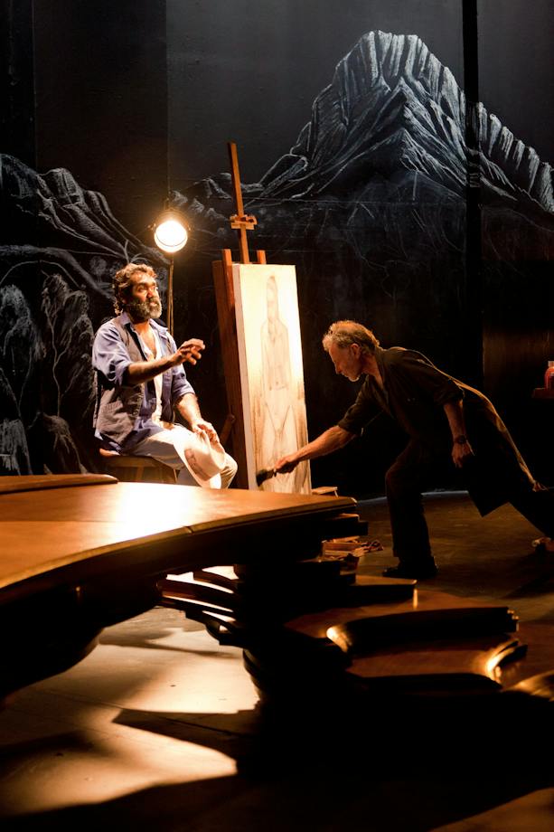 Two people on a darkened stage, one seated beside an easel while the other paints a portrait.