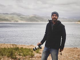 Photographer, Nathan White, posed with his camera in front of a lake lined with mountains.