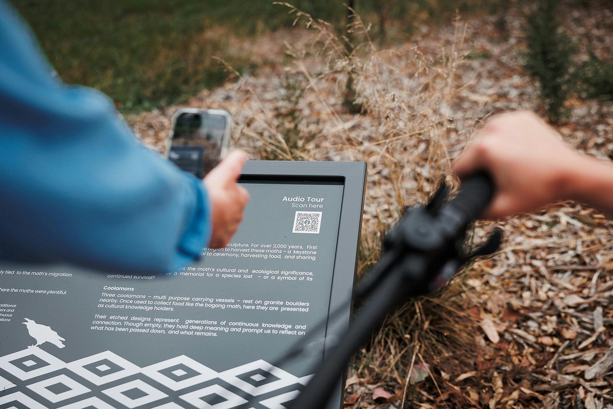 A person scanning a QR code on a black and white sign reading 'Audio Tour' with Indigenous patterns