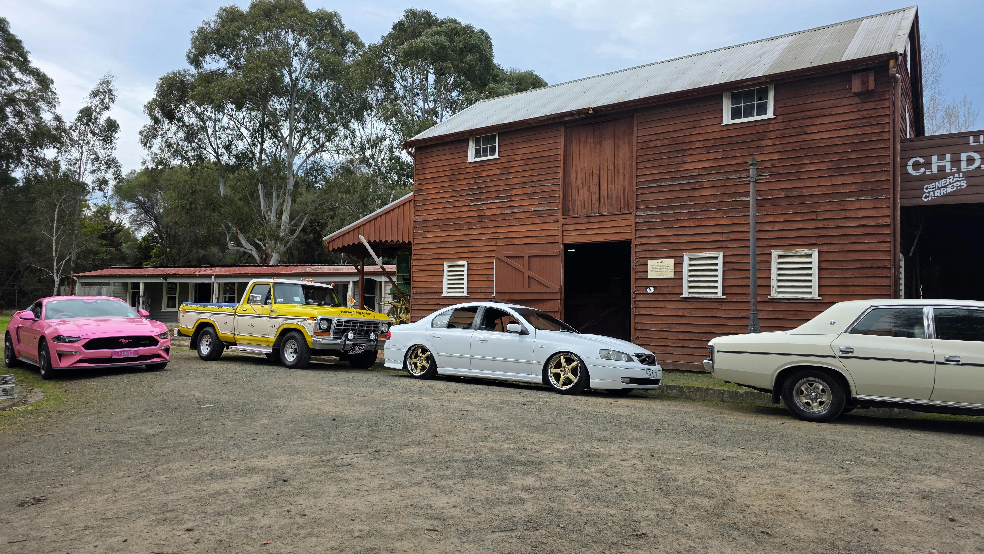 Cars of varying age parked alongside an old, two-story stable