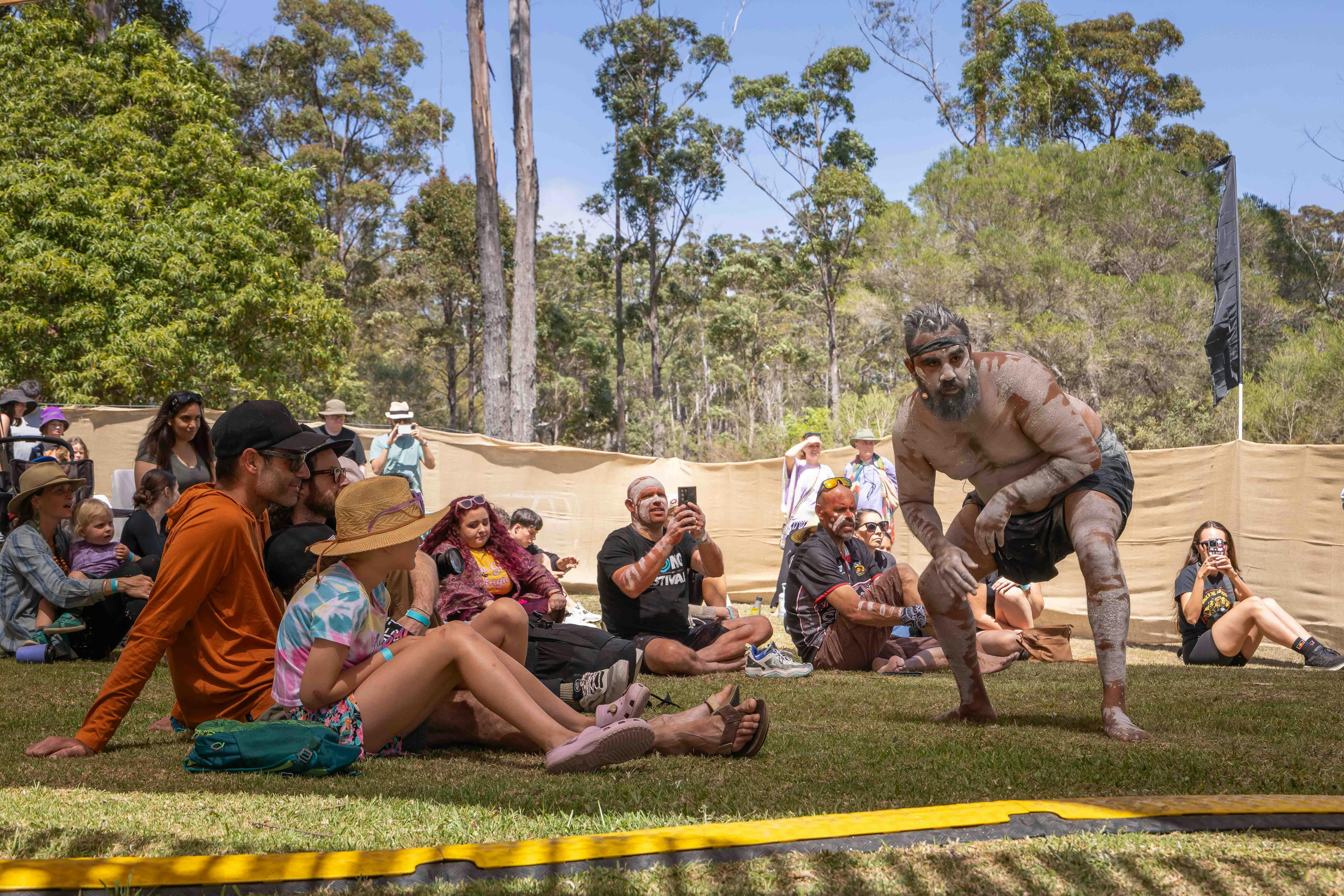 An Aboriginal male dancer poses for audience seated on the grass.