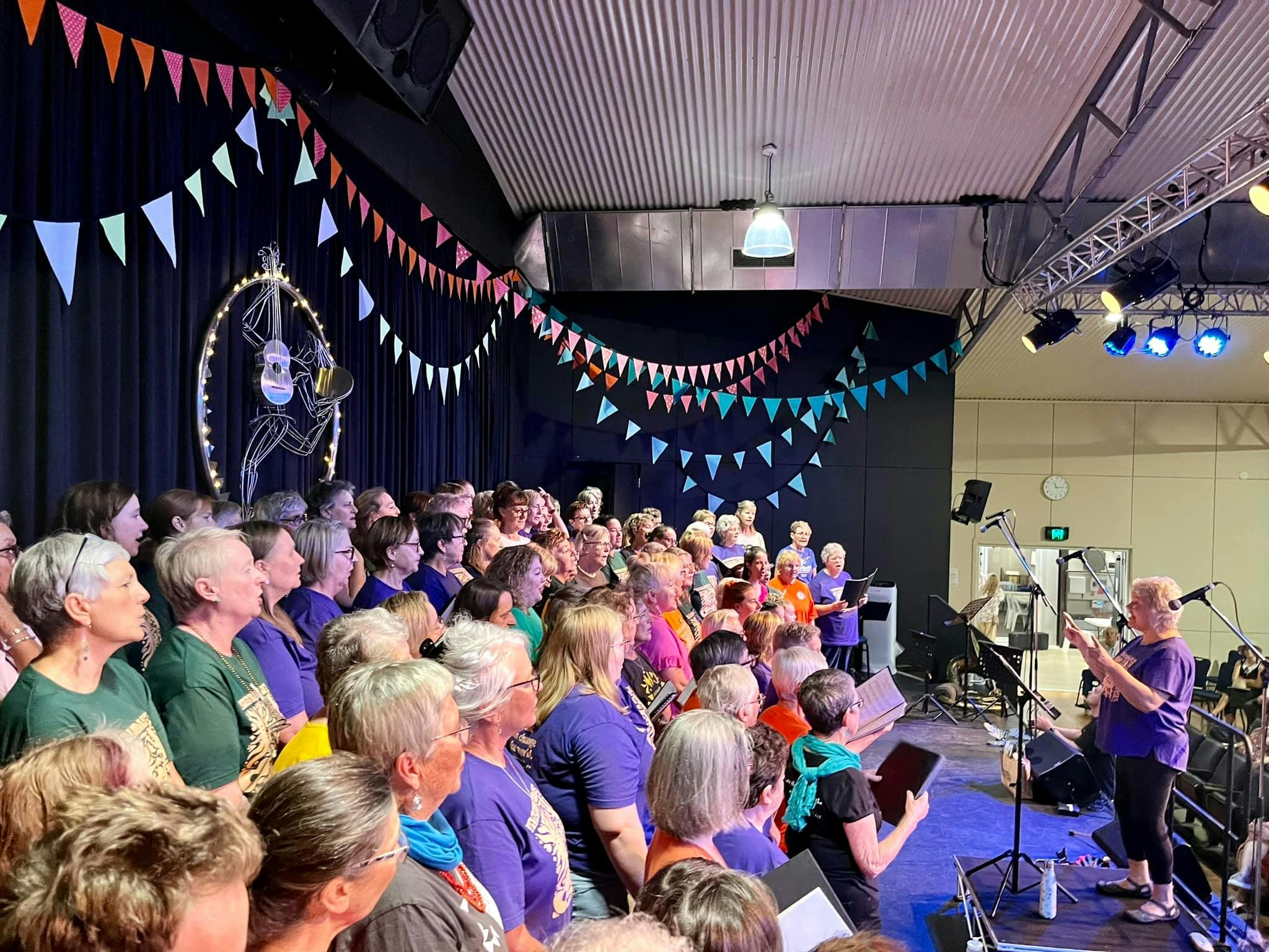 Approximately 60 choir members wearing colourful clothes singing on a large stage