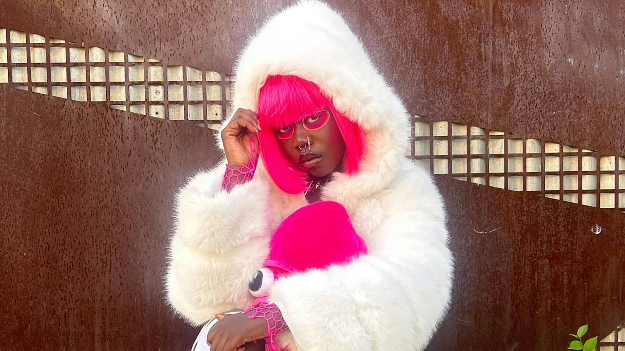 A woman with bright pink hair in a white fur coat posing in front of a rusted wall