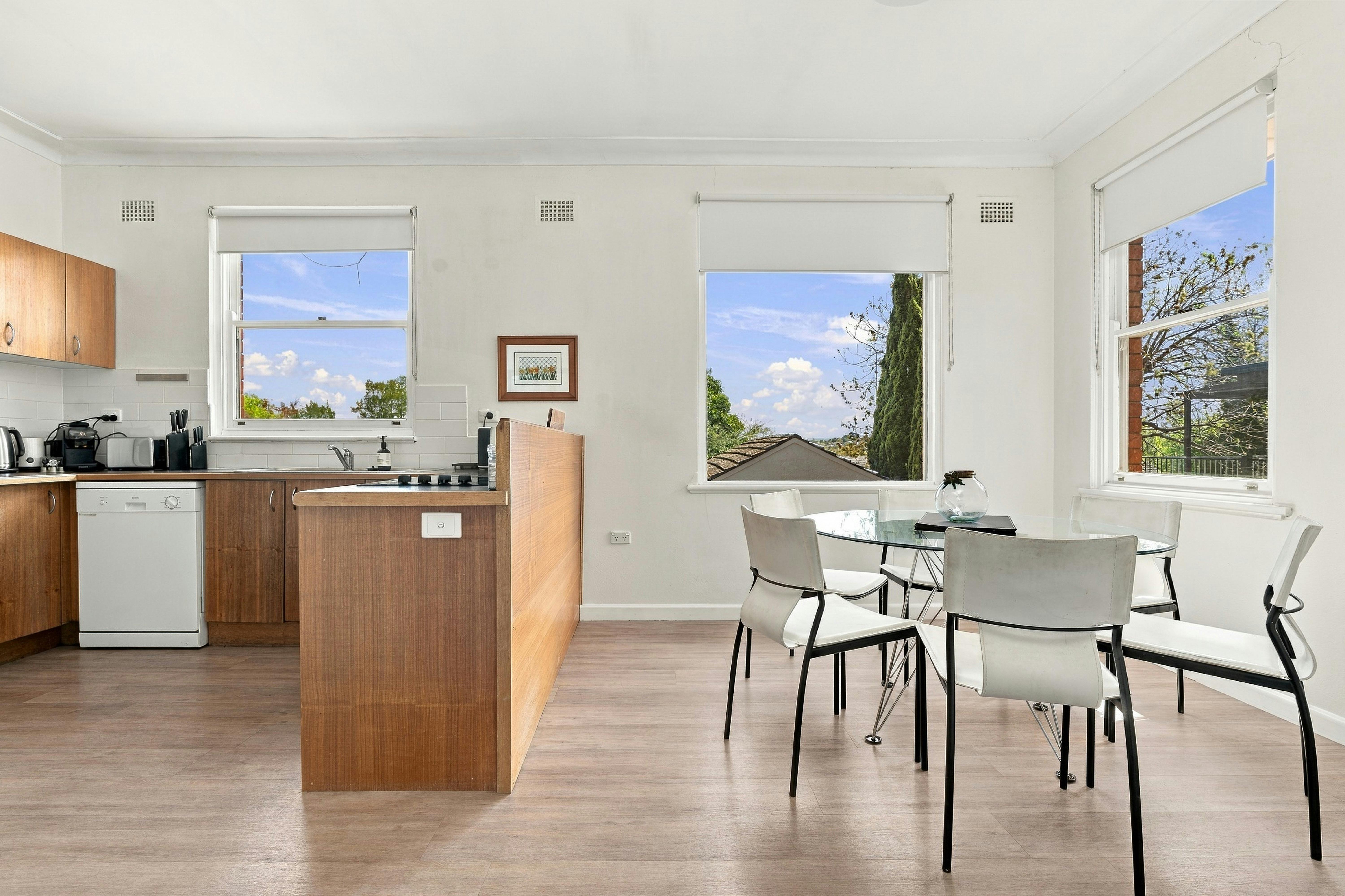 glass dining table, white dining chairs, large windows and wooden kitchen