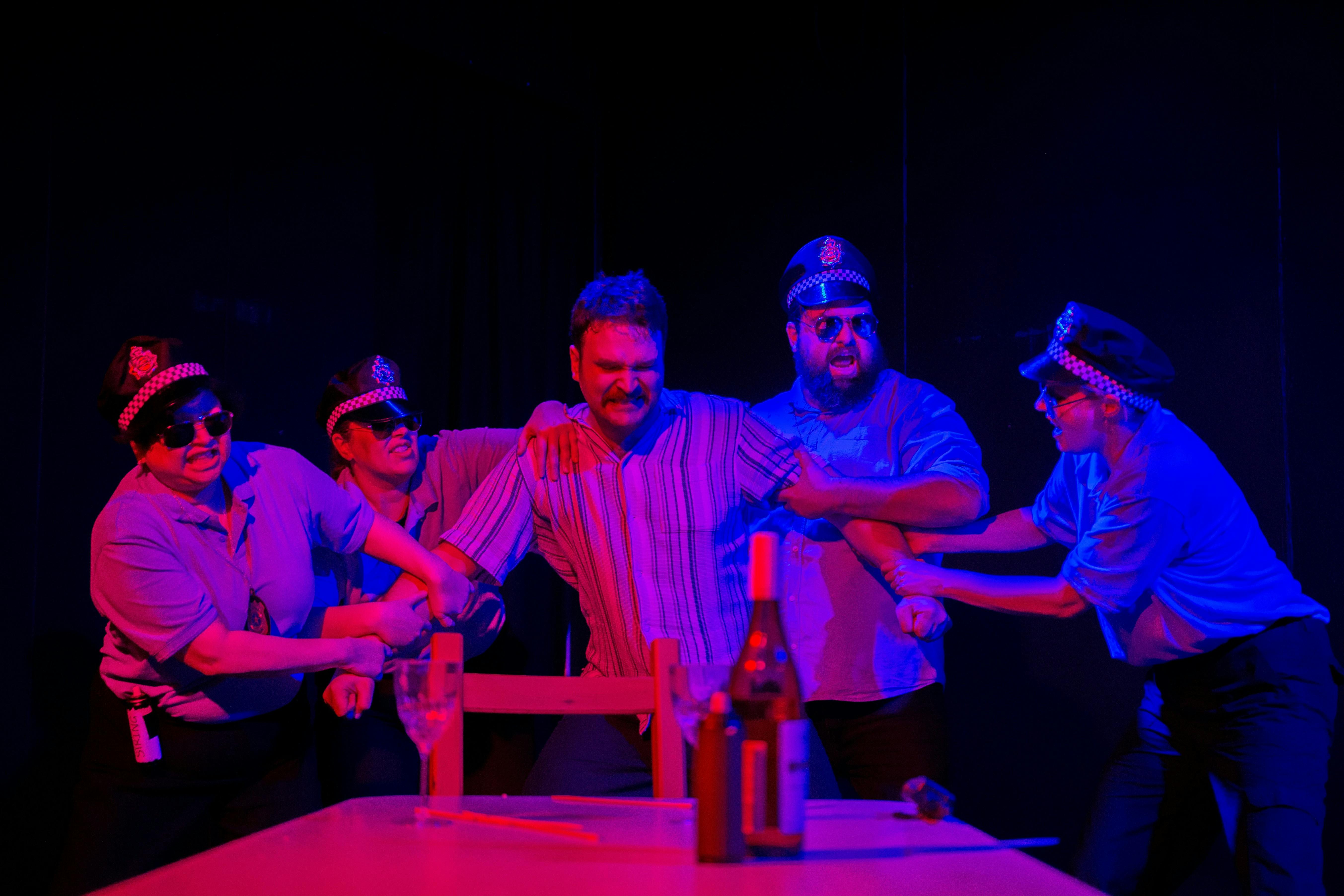 A man in a striped shirt, his arms held by police officers around him. In front, a table with wine.