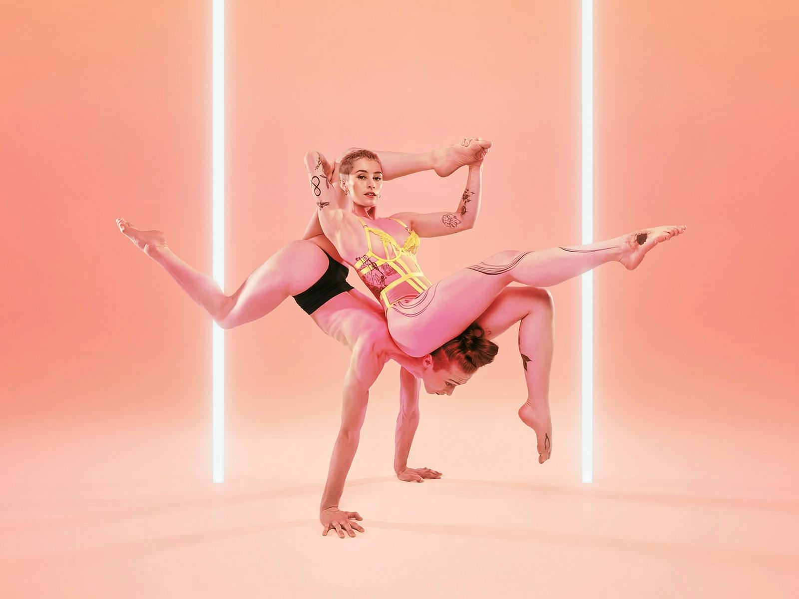 Two female acrobats hold a striking pose, one is in a handstand and the other is sitting on her