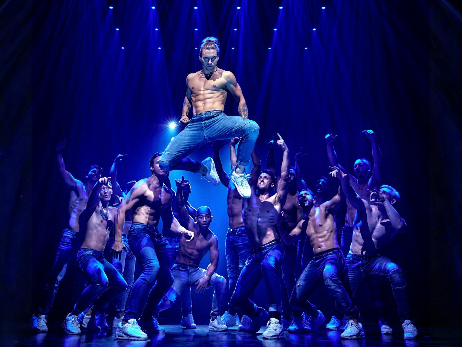 A shirtless dancer in jeans leaps above a crowd of bare-chested performers reaching upward.