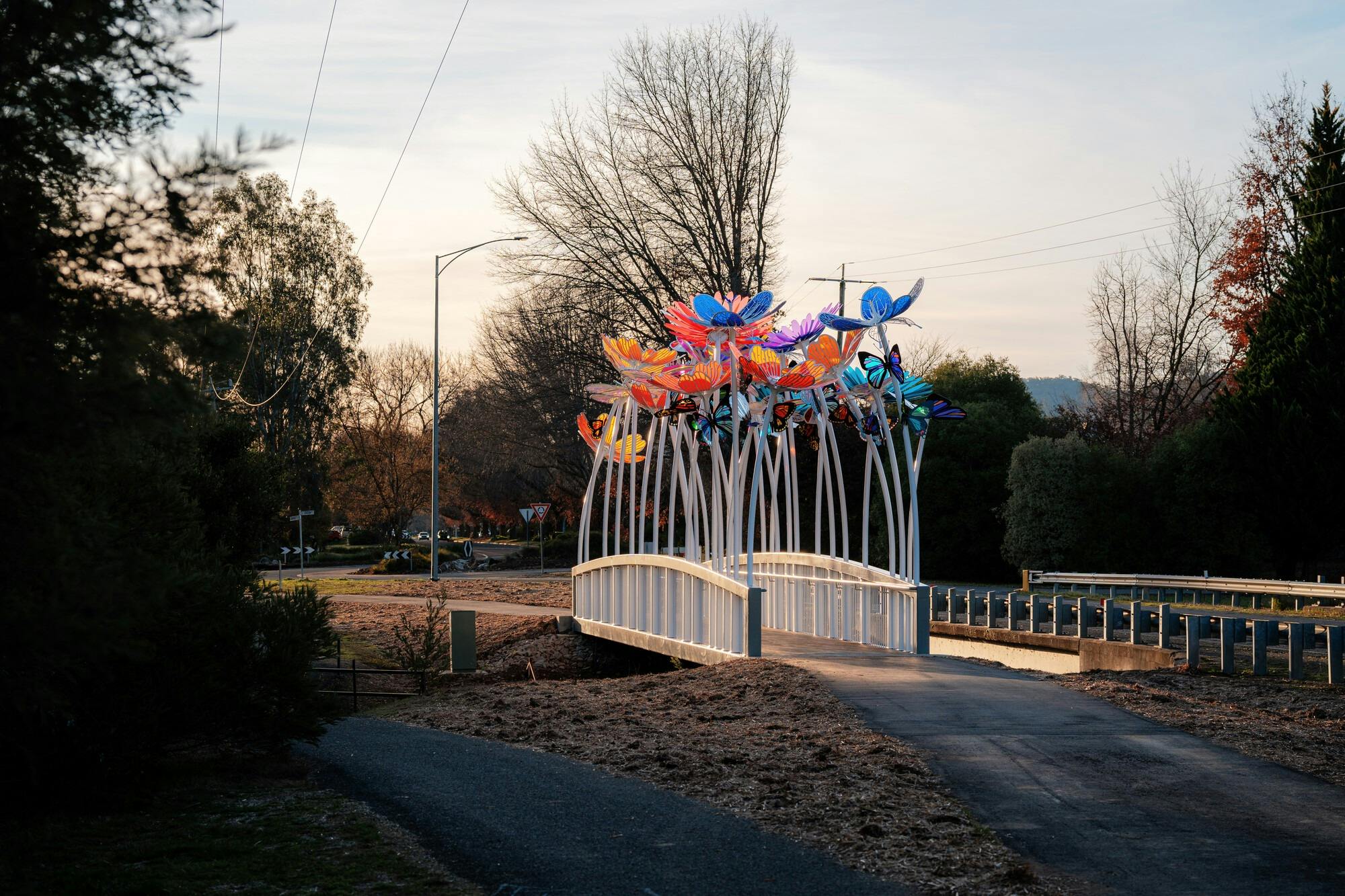 Two cyclists riding through a white, narrow bridge with tall colourful flower sculptures along it