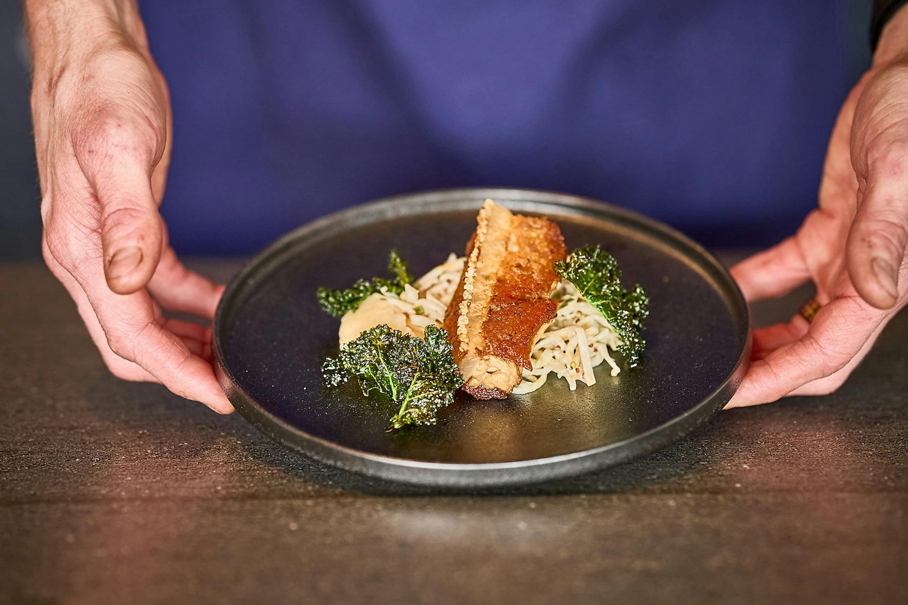Chef holding plate, presenting pork belly, celeriac remoulade, apple & cavolo nero