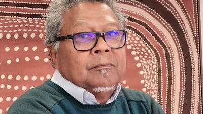 Portrait of an older individual wearing glasses and a dark green sweater, standing with arms crossed