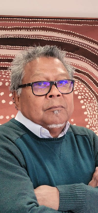 Portrait of an older individual wearing glasses and a dark green sweater, standing with arms crossed