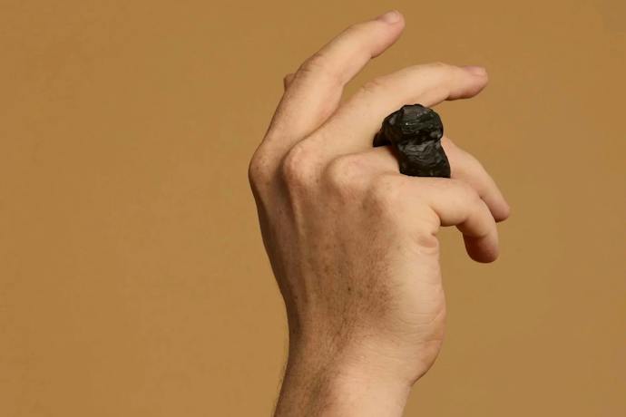 A hand wears a large ring carved out of a dark material against a tan background