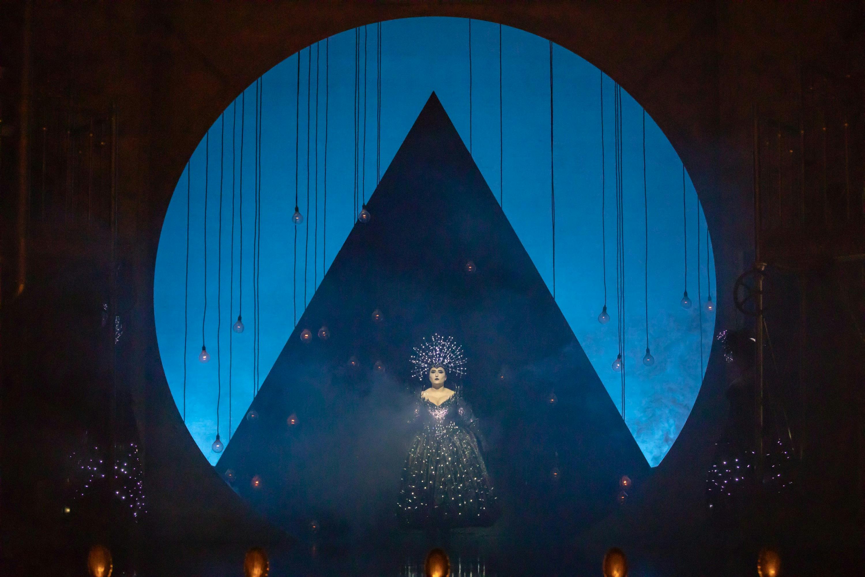 Performer in a sparkling costume stands before a blue-lit circle and pyramid on a dramatic stage.