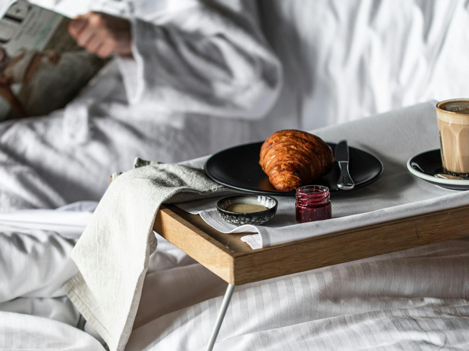 A person sitting in the bed with a tray with a croissant and a coffee