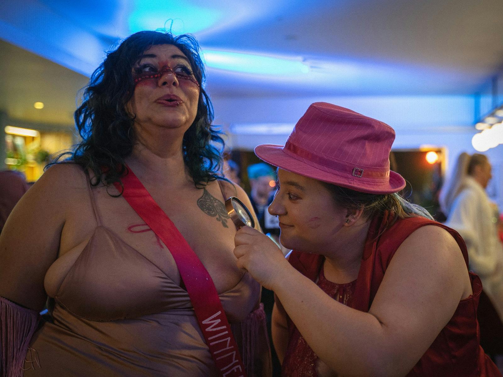 two AFAB performers in pink. one examines the other's chest with a magnifying glass